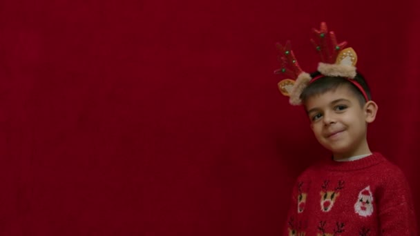 Lindo niño agitando la cabeza con orejas de reno y cuernos. Aislado en rojo. Feliz navidad. Cámara lenta con espacio de copia. Imágenes de alta calidad 4k - Imágenes, Vídeo