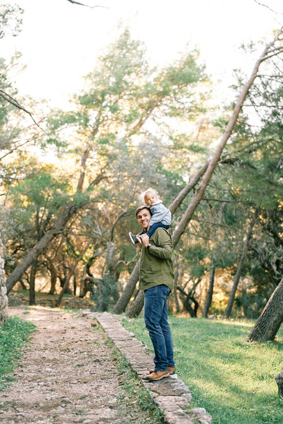 Uśmiechnięty tata z małą dziewczynką na ramionach stoi w połowie skręcony na utwardzonej ścieżce w lesie. Wysokiej jakości zdjęcie - Zdjęcie, obraz