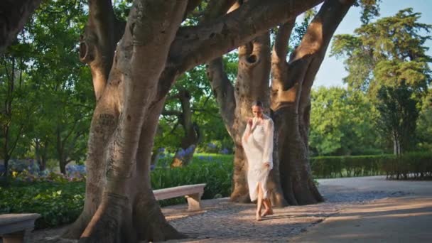 Nyugodt hölgy élvezi a sétát a gyönyörű zöld parkban. Romantikus fiatal nő sétál napfényes kertben fehér ruhát fújt a szél. Gyönyörű elegáns lány pihenni egyedül megy sikátorban hétvégén. - Felvétel, videó
