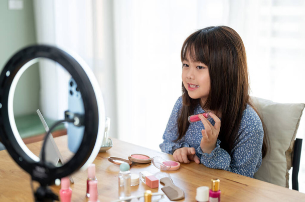 Een getalenteerd en schattig jong Aziatisch meisje beoordeelt haar kind cosmetische op haar sociale media, live streaming online thuis. Kid en technologie concepten, Kid schoonheid blogger - Foto, afbeelding