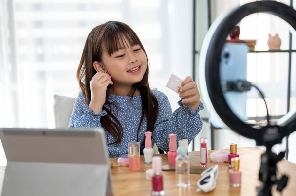 Una adorable joven influencer asiática está usando pendientes, grabando su tutorial de maquillaje para niños y preparándose para video o transmisión en vivo en sus redes sociales. Conceptos de niño y tecnología - Foto, imagen