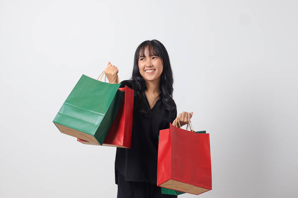 Portrait of excited Asian woman in black shirt holding and showing colorful paper shopping bags. Shopaholic girl and discount buying concept. Isolated image on white background - Photo, Image