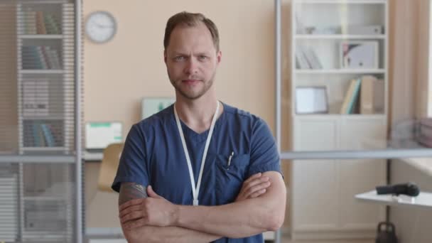 Retrato de fisioterapeuta masculino grave en uniforme médico de pie en la clínica y posando para la cámara con los brazos cruzados - Metraje, vídeo