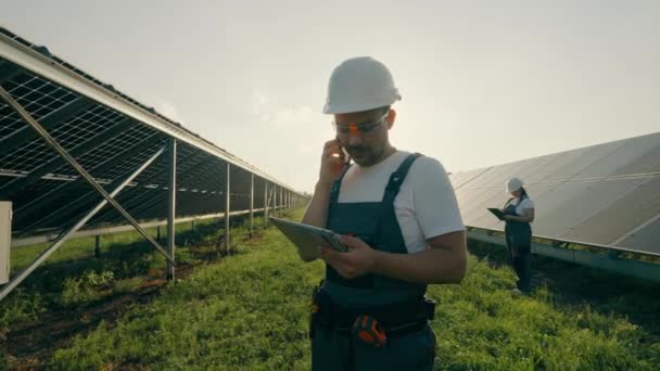 Ritratto di un operaio che parla al telefono vicino a una centrale solare e trasmette dati da un blocco note. Efficienza delle centrali solari. Scatto di alta qualità in formato 4K - Filmati, video