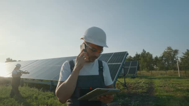 太陽光発電所に立っている間,携帯電話で話している監督エンジニアの肖像画. バックグラウンドでソーラーパネルを検査する労働者. 高品質の4K映像 - 映像、動画