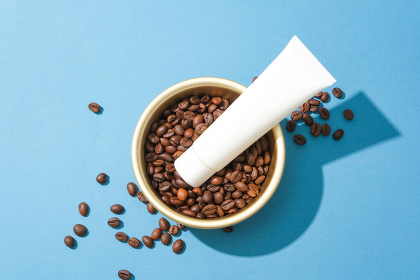 Detailní záběr kosmetické trubice umístěné na kávových zrnech v keramické misce. Modré pozadí pro reklamu. Káva má antibakteriální vlastnosti, které pomáhají omezit akné na kůži. - Fotografie, Obrázek