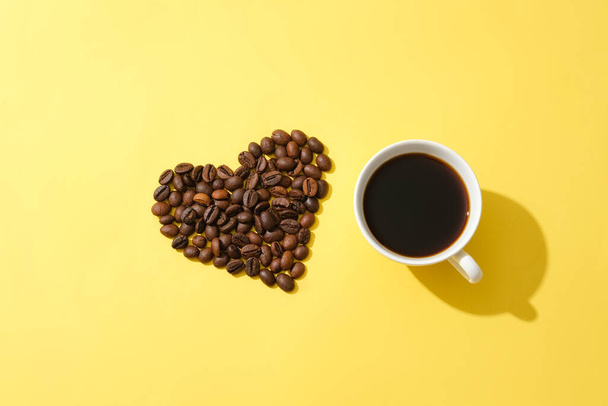 Kaffeebohnen in Herzform und eine Tasse schwarzen Kaffees auf gelbem Hintergrund. Die Anwendung einer Kaffeemaske hilft Sommersprossen zu verblassen und winzige Falten im Zusammenhang mit dem Altern abzuwehren. - Foto, Bild