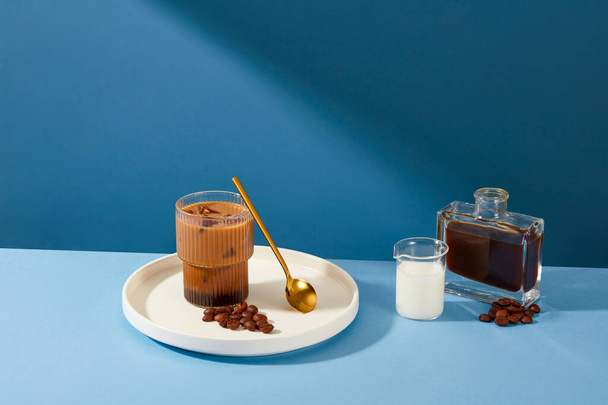 Eine Tasse Milchkaffee steht auf einem Keramikteller mit einem Metalllöffel und Kaffeebohnen, einer Tasse Milch und einer Flasche schwarzen Kaffees, die sich vor dem blauen Hintergrund abhebt.. - Foto, Bild