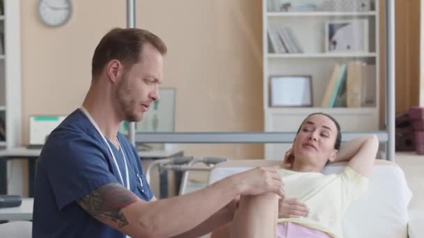 Физиотерапевт осматривает колено пациентки и рассказывает ей о плане лечения в клинике - Кадры, видео