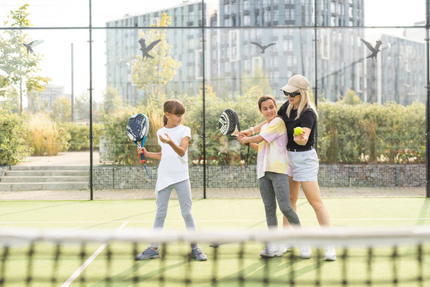 Mladý tenisový trenér v aktivním oblečení drží raketu v držení jedné ze dvou roztomilých dívek při konzultacích s ní během individuálního tréninku na stadionu. Kvalitní fotografie - Fotografie, Obrázek