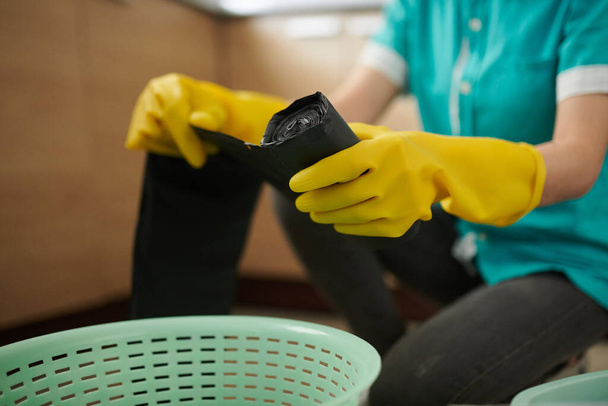 Gros plan de la travailleuse de nettoyage dans des gants de protection changeant de sac poubelle dans un seau pendant son travail dans la cuisine - Photo, image