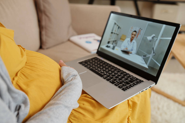 Крупный план беременной девушки, сидящей на диване с ноутбуком и разговаривающей с врачом во время онлайн-консультации - Фото, изображение