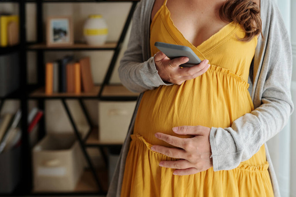 Primer plano de la mujer embarazada acariciando su vientre embarazada y deslizando la pantalla de su teléfono inteligente - Foto, imagen