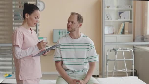 Γυναίκα φυσιοθεραπεύτρια σε scrubs κρατώντας πρόχειρο και έχοντας συνομιλία με αρσενικό ασθενή κατά τη διάρκεια της διαβούλευσης στην κλινική - Πλάνα, βίντεο