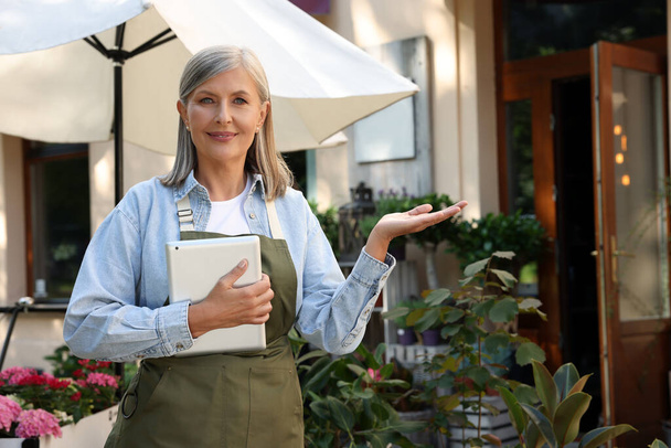 Happy business owner with tablet inviting to come into her flower shop outdoors, space for text - Photo, image