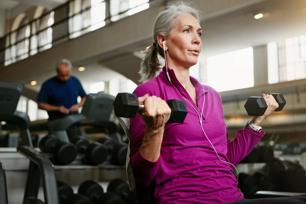Dumbbell, fitness and senior woman at a gym for training, wellness and cardio with earphones, music or mindset. Weightlifting, bodybuilding and elderly female person at sports center for arm workout. - Photo, Image