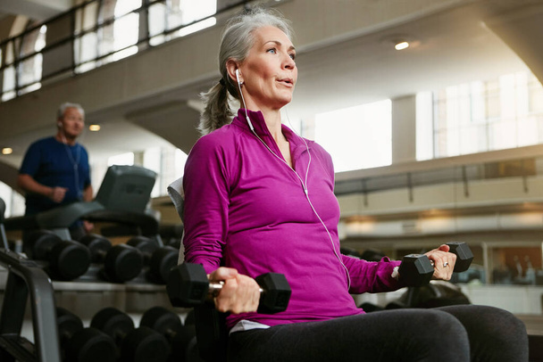 Fitness, dumbbell and senior woman at a gym for training, wellness and cardio with earphones, music or mindset. Weightlifting, bodybuilding and elderly female person at sports center for arm workout. - Photo, Image