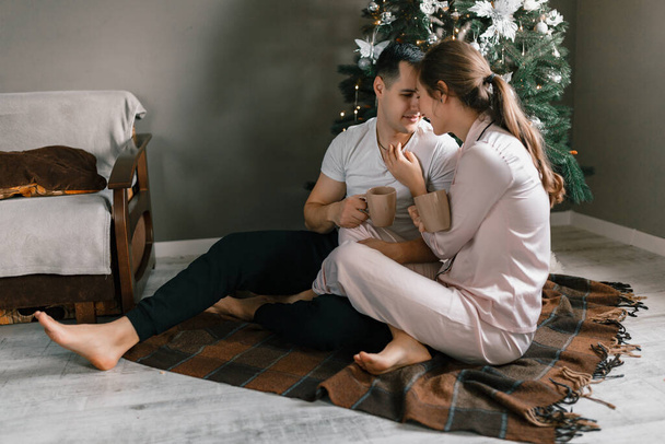 Una joven y feliz pareja abrazándose mientras están sentados junto al árbol de Navidad en casa. Una hermosa mujer y un hombre en pijama relajarse en su apartamento - Foto, imagen