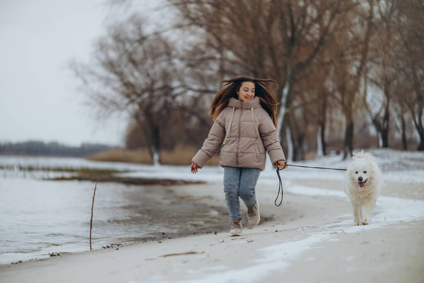 Ein Mädchen spaziert mit ihrem geliebten Haustier Samoyed im Winter am Ufer eines Sees im Park. Im Winter mit dem Hund spazieren. - Foto, Bild