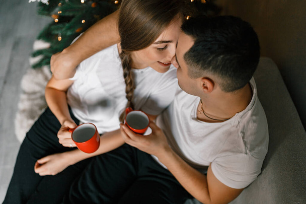 Una pareja joven y alegre está sentada en un cómodo sofá cerca del árbol de Navidad en su casa. El atractivo hombre y la mujer se deleitan en el sorbo de chocolate caliente, creando un ambiente acogedor en el hogar durante las vacaciones de Año Nuevo - Foto, Imagen