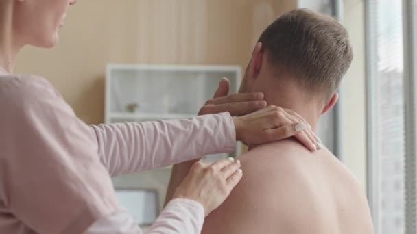 Fizjoterapeutka dotykająca szyi i ramienia pacjenta bez koszulki, podczas diagnozowania bólu mięśni podczas konsultacji lekarskiej w klinice - Materiał filmowy, wideo
