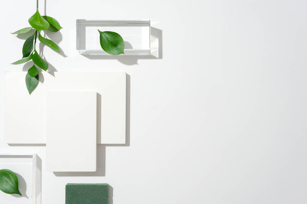 白い背景の左側,2つのガラスプラットフォーム,白い表彰台,緑の葉がダイナミックな構成を作成します. 魅力的な商業ビジュアルのための十分な空スペースでテキストを設計する. - 写真・画像