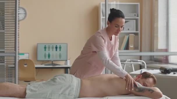 Fisioterapeuta femenina que da masaje de espalda a un paciente masculino acostado en un sofá médico en una clínica de bienestar - Imágenes, Vídeo