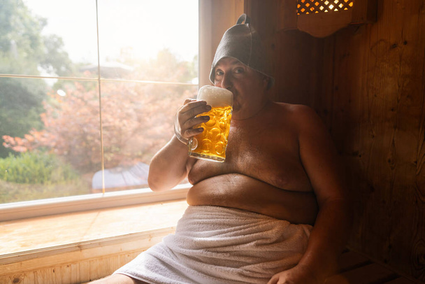 Mies suomalaisessa saunassa huopahattu päässä, siemaillen saksalaista olutmukia, kirkas ikkuna taustalla. Oktoberfestin konseptikuva - Valokuva, kuva