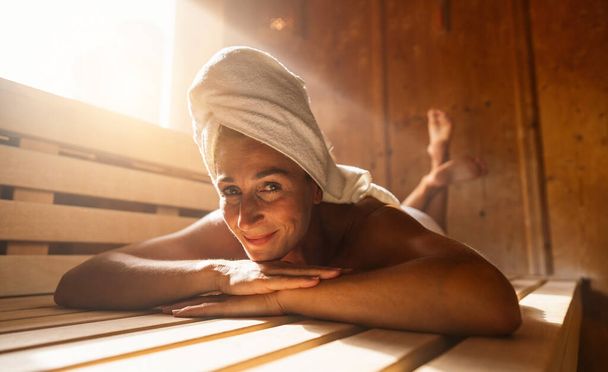 Donna che si rilassa in una sauna finlandese, con un asciugamano in testa, sorridendo alla telecamera - Foto, immagini