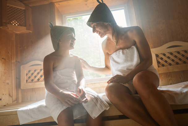 Madre e hijo sonriéndose en una sauna finlandesa, ambos llevando sombreros de fieltro - Foto, imagen