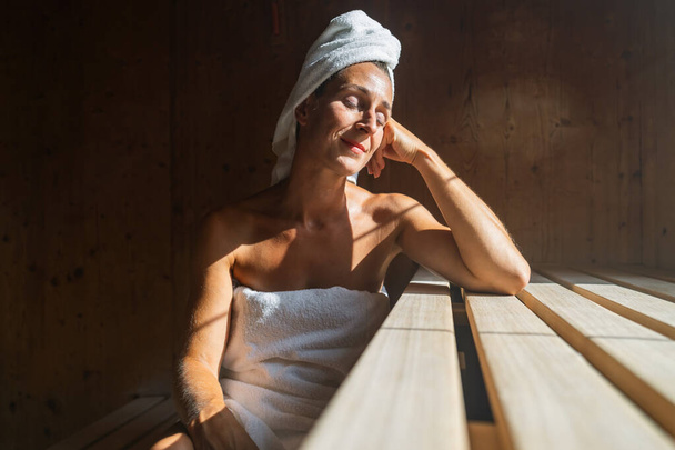 Mujer en una sauna finlandesa inclinada hacia atrás, con los ojos cerrados, con una toalla envuelta en su cabeza, iluminada por el sol en un balneario - Foto, imagen