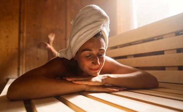 Donna contenta sdraiata su una panchina finlandese con un asciugamano avvolto intorno alla testa. Spa wellness hotel concetto immagine. - Foto, immagini