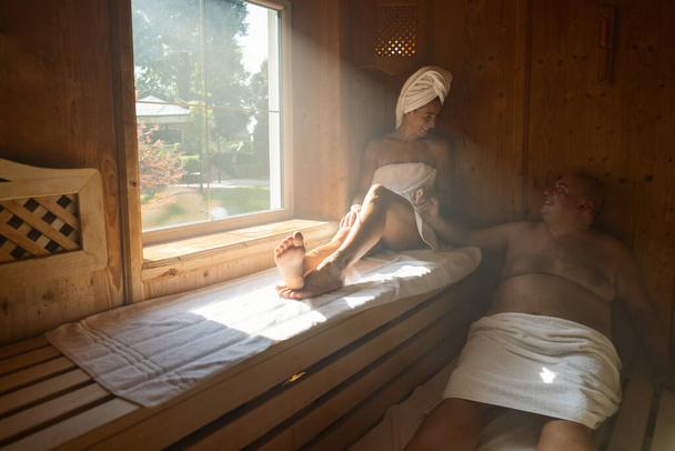 Couple relaxing in a sauna, woman with a towel on her head, both seated and smiling at spa hotel - Photo, Image