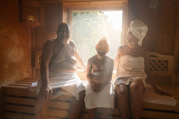 Family in a finnish sauna laughing together, man on left, woman with towel on head on right, child in middle at wellness hotel - Photo, Image
