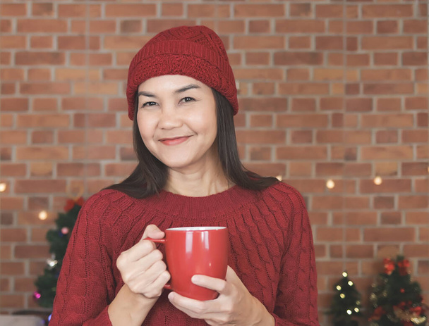 Πορτρέτο της Ασίας γυναίκα φορώντας κόκκινο πλεκτό πουλόβερ και καπέλο στέκεται στην κουζίνα με χριστουγεννιάτικες διακοσμήσεις, πίνοντας καφέ από την κόκκινη κούπα, χαμογελώντας ευτυχισμένοι και κοιτάζοντας κάμερα. χειμώνας και Χριστούγεννα. - Φωτογραφία, εικόνα