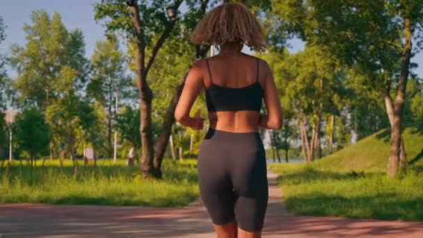 задний вид женщина одета шорты и топ бег трусцой в городском парке кардио тренировки летом замедленного движения - Кадры, видео