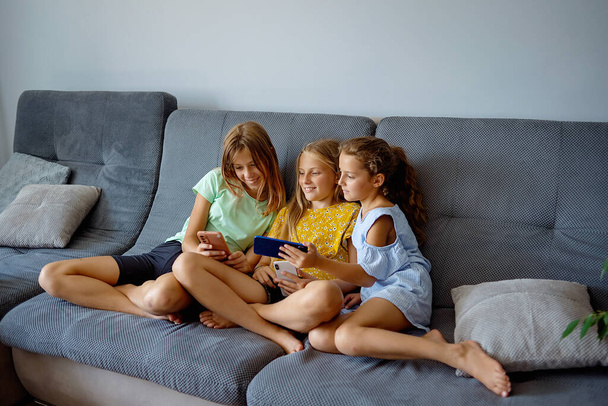 Девушки играют в мобильную игру на смартфоне, сидя на диване. Детский досуг дома, зависимость от видеоигр - Фото, изображение
