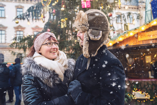 Kış kıyafetleri içinde gülümseyen bir çift, karlı bir Noel pazarına yakın duruyorlar, arkalarında da neşeli ışıklar var.. - Fotoğraf, Görsel