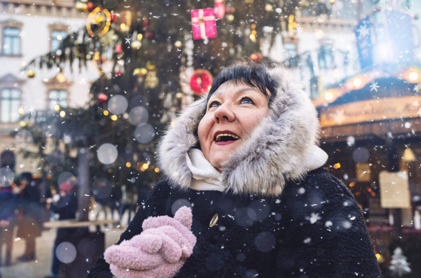Seniorin im Pelzmantel schaut freudig auf, als auf dem Weihnachtsmarkt Schnee fällt - Foto, Bild