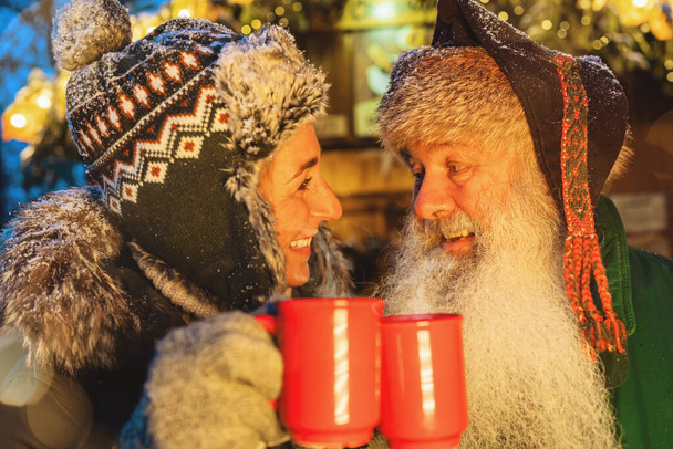 vrienden toasten met glühwein of warme chocolademokken op kerstmarkt, vrouw met wintermuts, man met witte baard - Foto, afbeelding