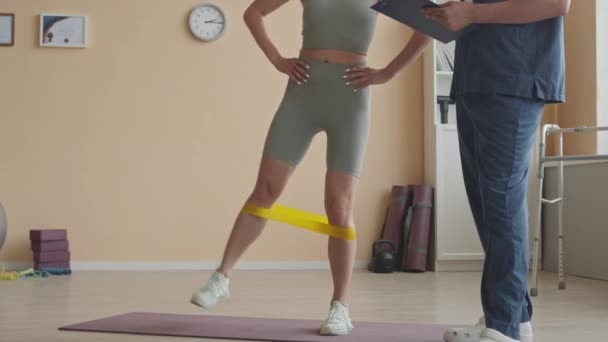 Vágott lövés női sportruházat csinál oldalsó láb emeli rezisztencia sávban, miközben a rehabilitációs terápia ülésén fizioterapeuta a klinikán - Felvétel, videó