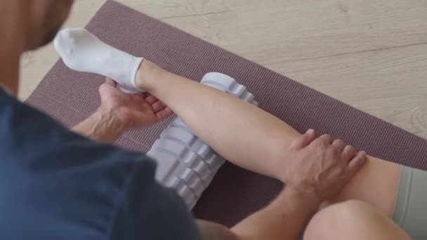 Přes rameno záběr profesionálního fyzioterapeuta pomáhá pacientce při masáži tele s pěnovým válečkem na rohožce během rehabilitačního sezení na klinice - Záběry, video