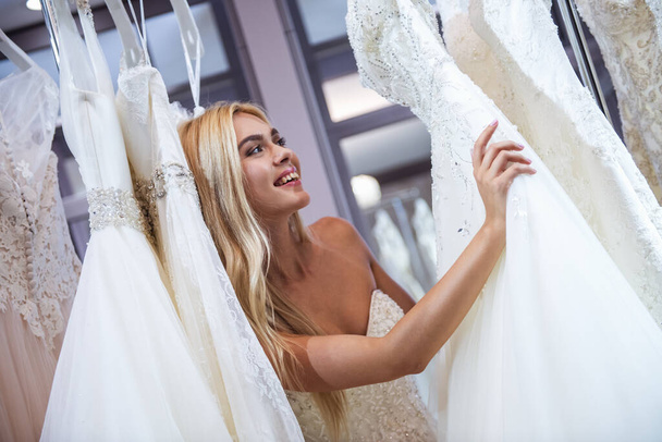 Attractive young bride is smiling while choosing wedding dress in modern wedding salon - Photo, Image