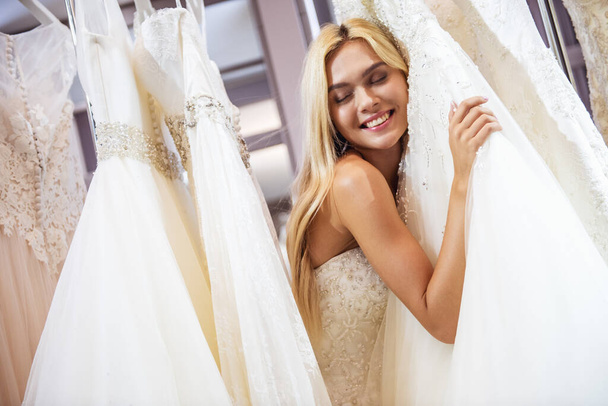 Atractiva novia joven está sonriendo al elegir el vestido de novia en el salón de bodas moderno
 - Foto, imagen