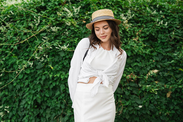 Романтичная брюнетка в соломенной шляпе, позирующая с застенчивой улыбкой. Грациозная молодая женщина летом на природе. Копирование пространства - Фото, изображение