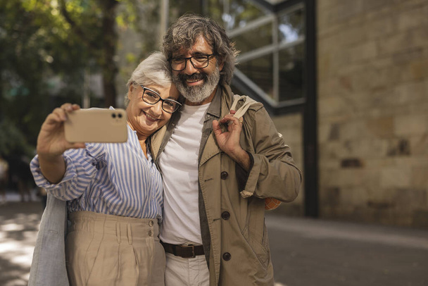 Cheerful mature couple using a smartphone to take a selfie in the middle of a street in the city with a sunny day and remembering their Valentine's date - Photo, Image