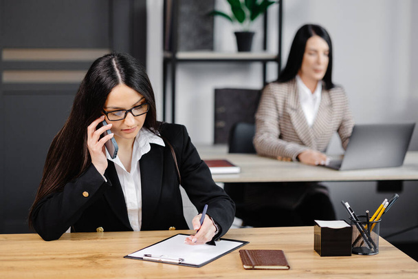 Atrakcyjna młoda kobieta biznesu w kurtce rozmawiająca przez telefon komórkowy, siedząca na biurku i korzystająca z notebooka obok kolegi z pracy w biurze. - Zdjęcie, obraz