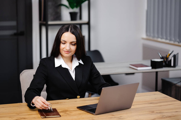 Νεαρή ευτυχισμένη υπάλληλος επιχειρηματίας φορώντας κοστούμι κάθεται στο γραφείο με φορητό υπολογιστή και δίνοντας το κλειδί στο γραφείο. - Φωτογραφία, εικόνα