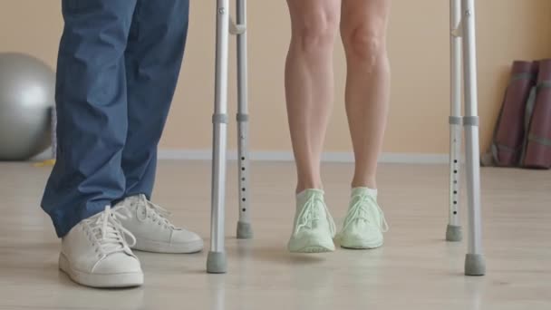 Colpo a sezione bassa delle gambe del fisioterapista che sostiene la paziente femminile utilizzando il telaio a piedi nella sessione di riabilitazione in clinica - Filmati, video