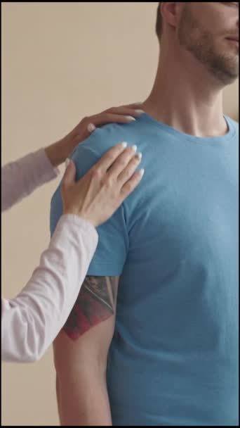 Вертикальный обрезанный снимок мужчины, стоящего в физиотерапевтической клинике и разговаривающего с женщиной-врачом, осматривающей его руку - Кадры, видео